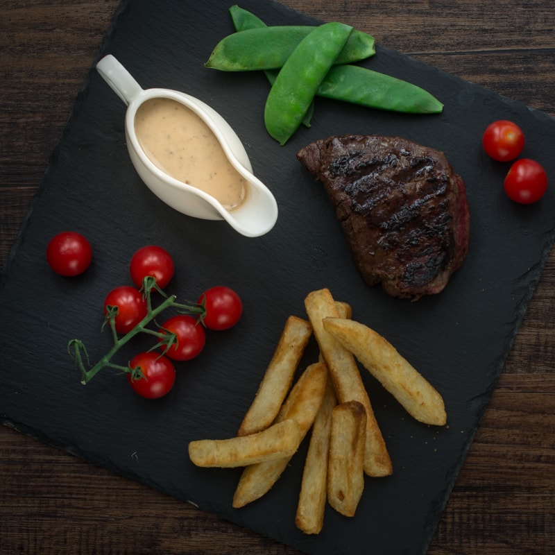 Looking down on a slate with a white jug of peppercorn sauce, cherry tomatoes, mangetout, chunky chips and steak.