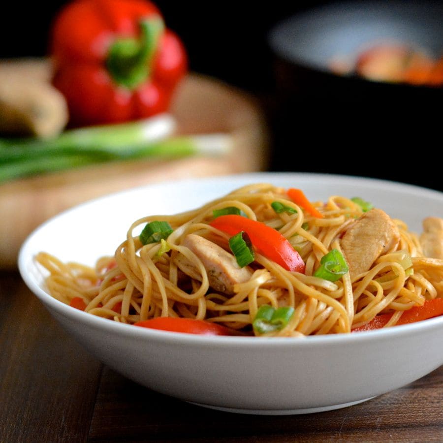 A bowl of homemade chicken chow mein with a wok and chopping board of ingredients in the background.
