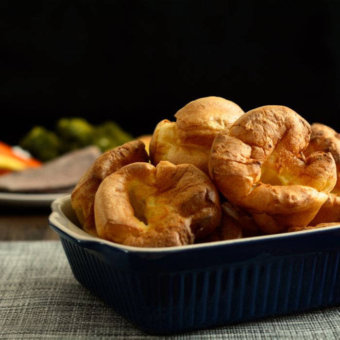 A blue serving dish full of freshly made Yorkshire puddings with a plate of roast dinner in the background.