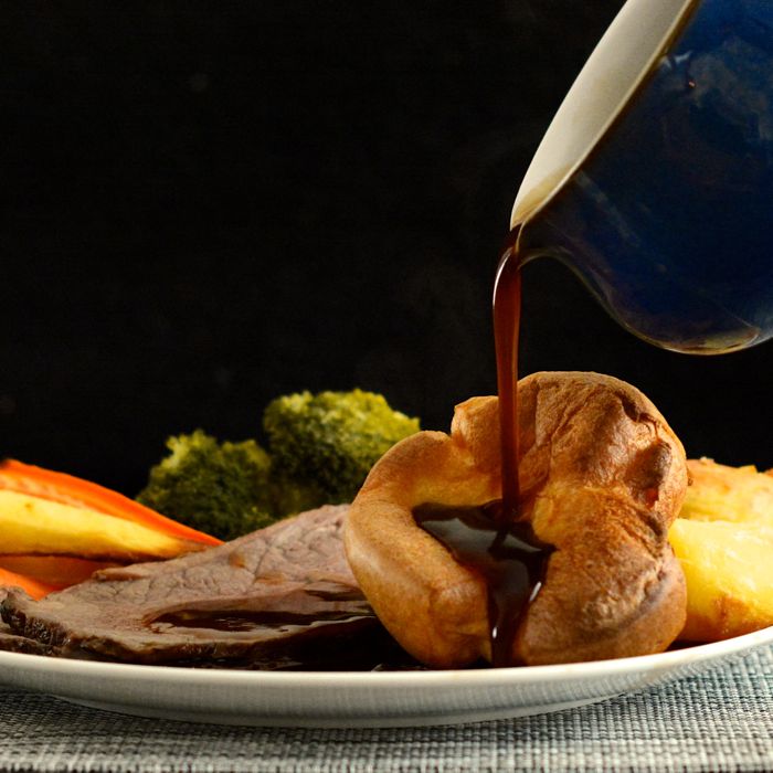 Gravy being poured onto a plate of roast beef with homemade Yorksire puddings.