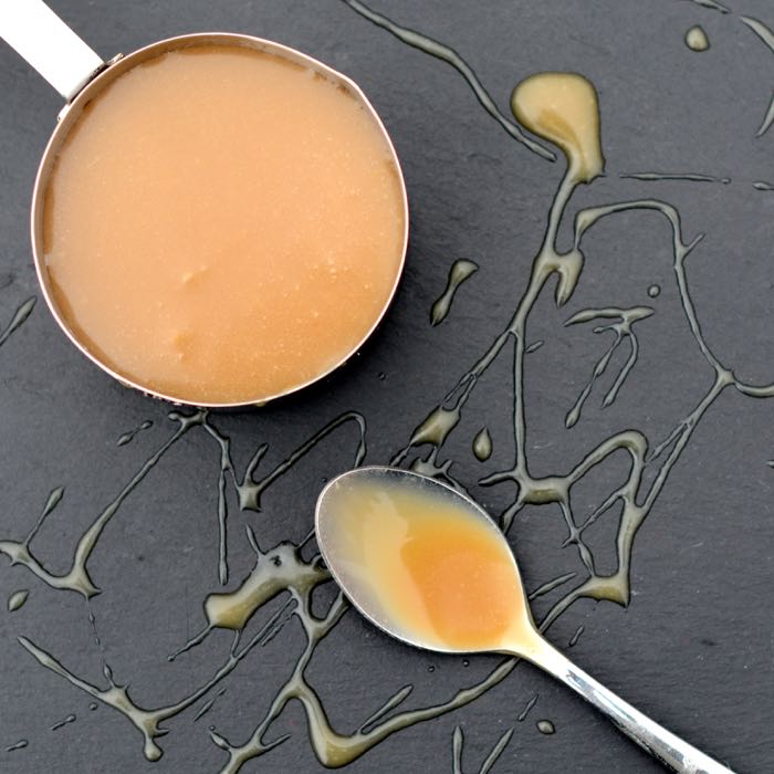 Looking down on a measuring cup full of caramel sauce with a caramel covered spoon next to it. This is all on a slate which has been drizzled with caramel sauce.