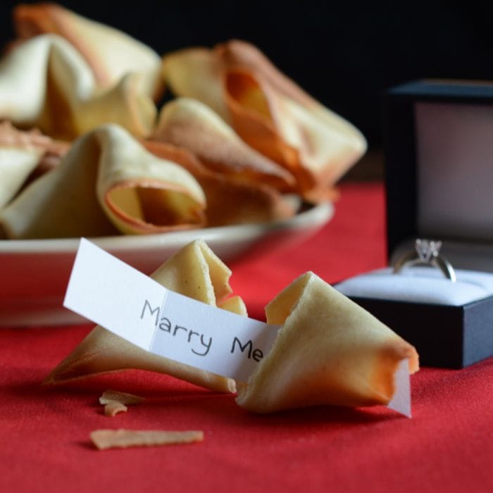 Close up of broken fortune cookie revealing message inside saying marry me with an engagement ring in the background. 