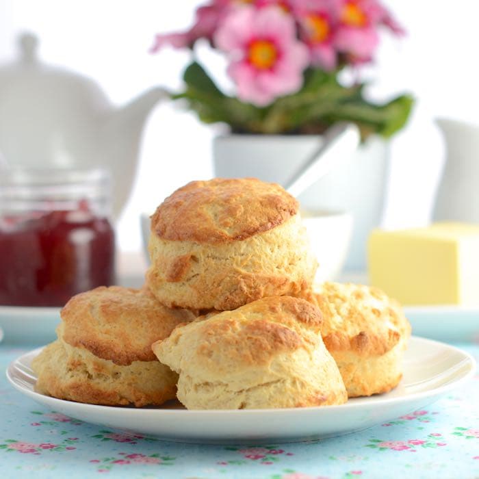 A stack of plain scones on a plate with tea, jam an flowers in the background.