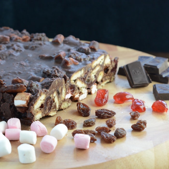 Rocky road shown with a slice removed so the inside fillings can be seem. Cherries, chocolate, marshmallows and raisins are scattered on the board. 