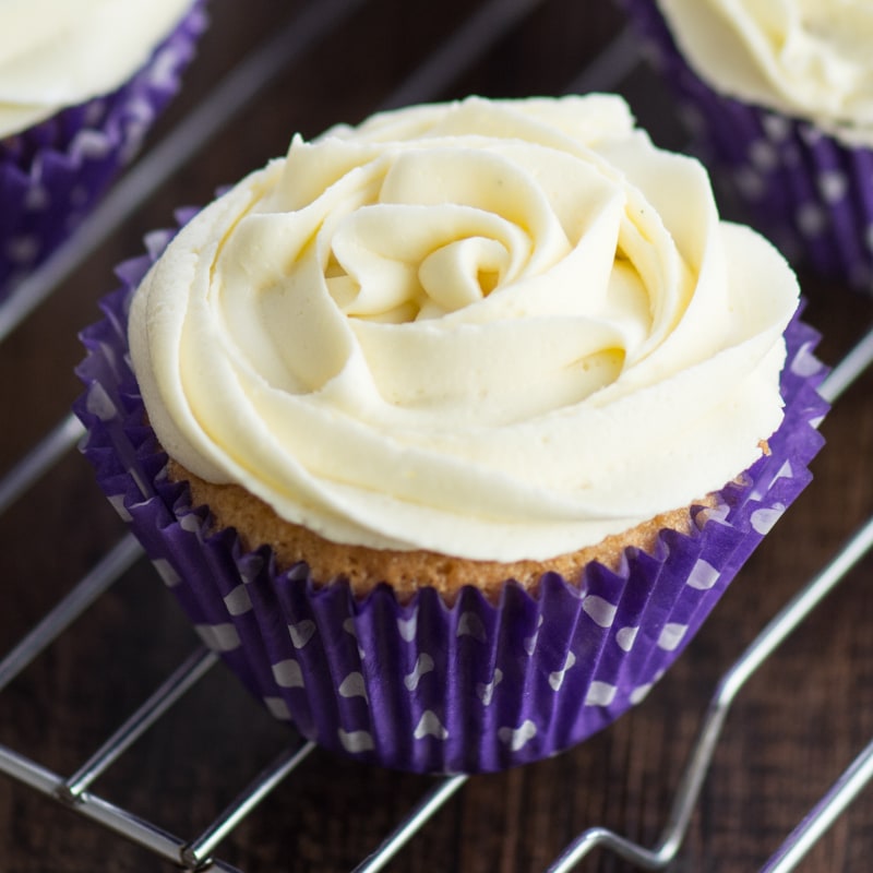 A close up of a vanilla cupcake topped with a vanilla buttercream rose in a purple cupcake case.