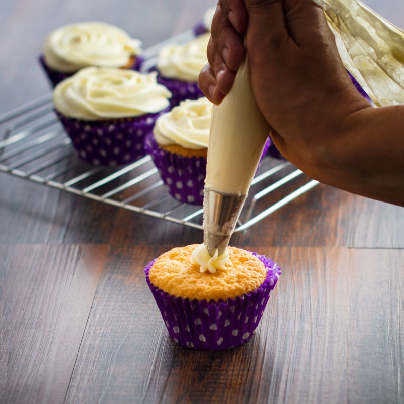 Vanilla buttercream being piped onto a cupcake with more iced cupcakes in the background.