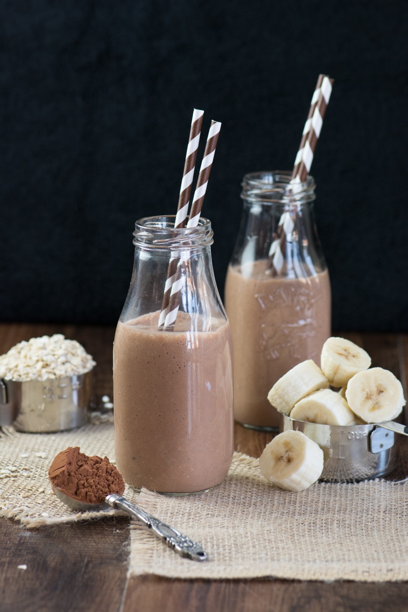 Two glass milk bottles filled with a coconut, banana & chocolate breakfast smoothie. Both have two brown and white striped straws and they're surrounded by some of the smoothie ingredients - banana, cocoa powder and oats.