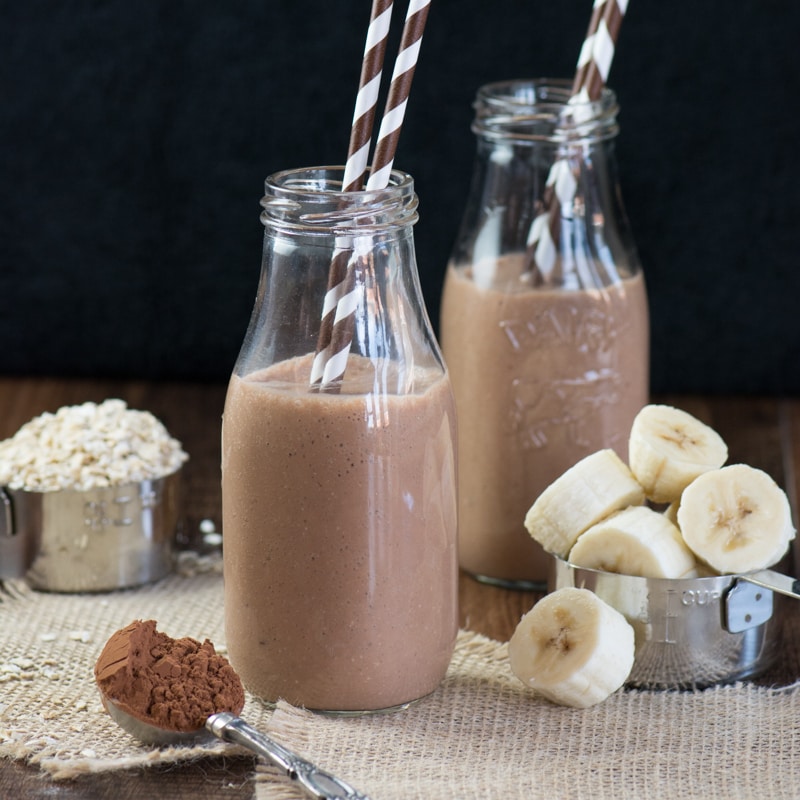 Two glass milk bottles filled with a coconut, banana & chocolate breakfast smoothie. Both have two brown and white striped straws and they're surrounded by some of the smoothie ingredients - banana, cocoa powder and oats.