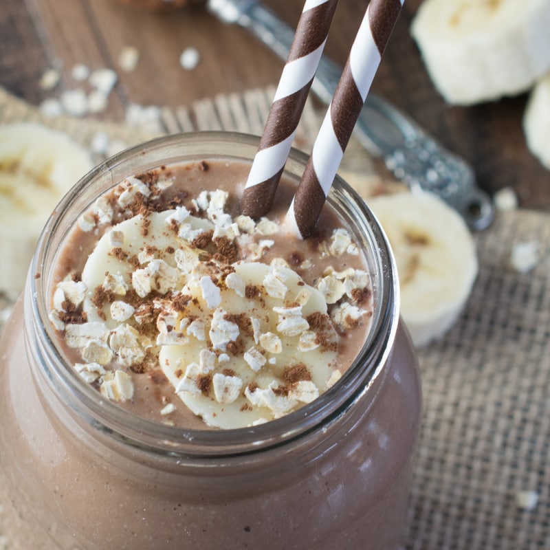 A close up of the coconut, banana & chocolate breakfast smoothie topped with oats, slices of banana and sprinkled with cocoa powder.