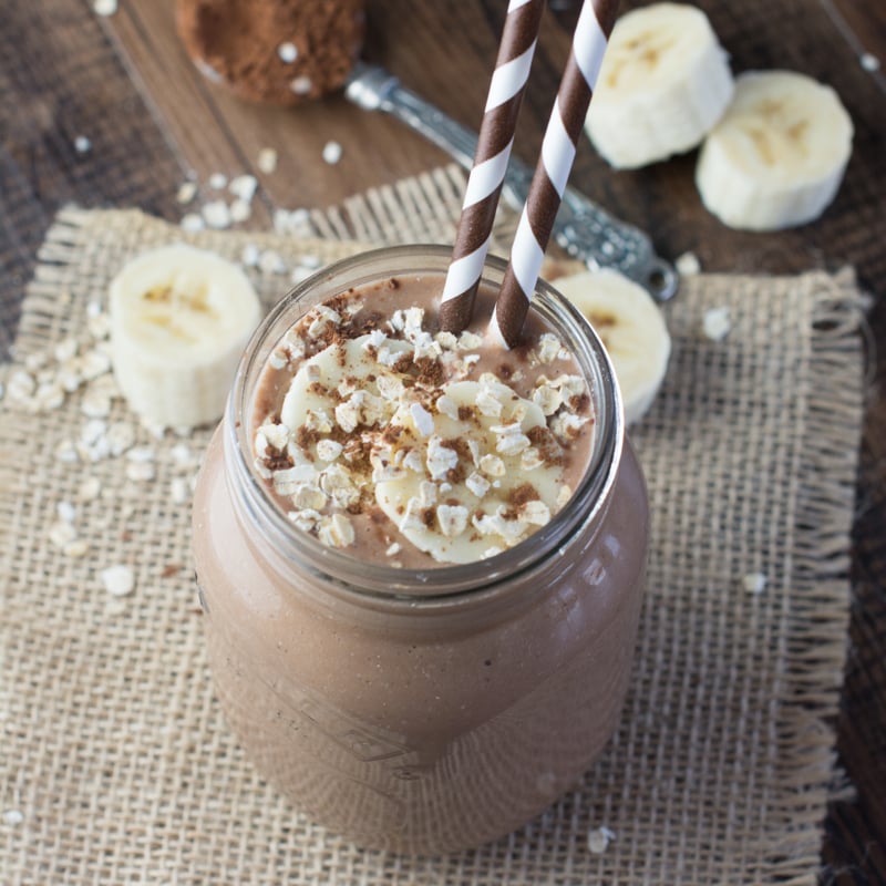 Coconut, Banana & Chocolate Breakfast Smoothie in a mason jar with two brown striped straws. The smoothie is topped with slices of banana and oats.