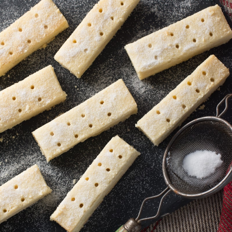 Shortbread cooling on a slate board shown from above with a small sieve of sugar. 