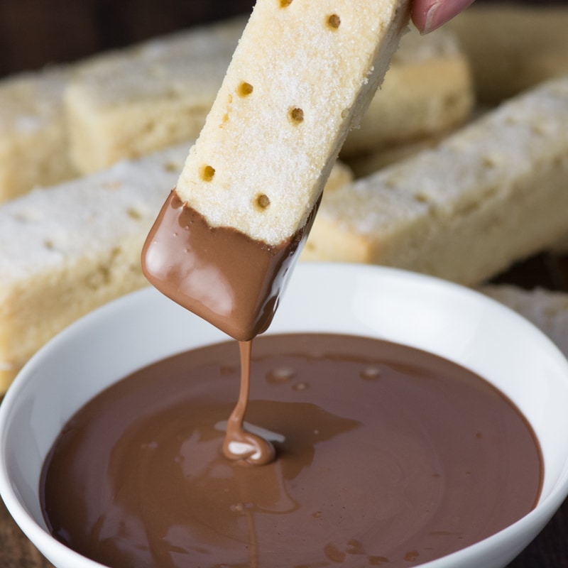 Close up of a shortbread finger that has just had the end dipped in a bowl of melted milk chocolate and is still dripping a trail of chocolate. 