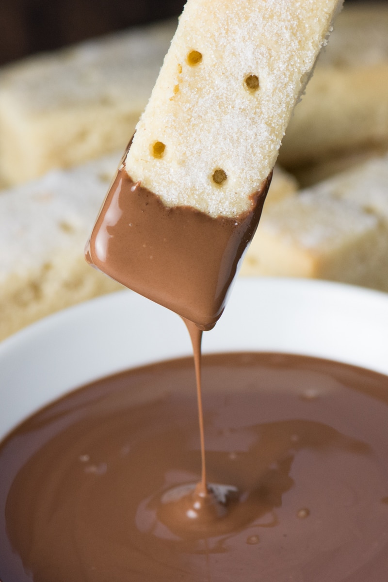 Extreme close up of a shortbread finger that has just had the end dipped in a bowl of melted milk chocolate and is still dripping a long trail of chocolate. 