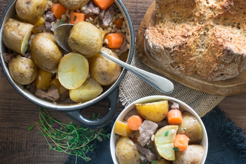 Overhead view of irish stew being served into one bowl with a spoon alongside a loaf of soda bread on a wooden board. 