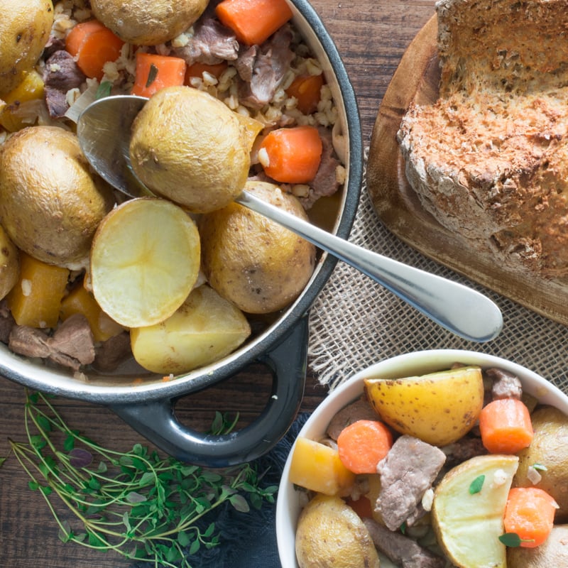Overhead view of irish stew being served into one bowl with a spoon alongside a loaf of soda bread on a wooden board. 