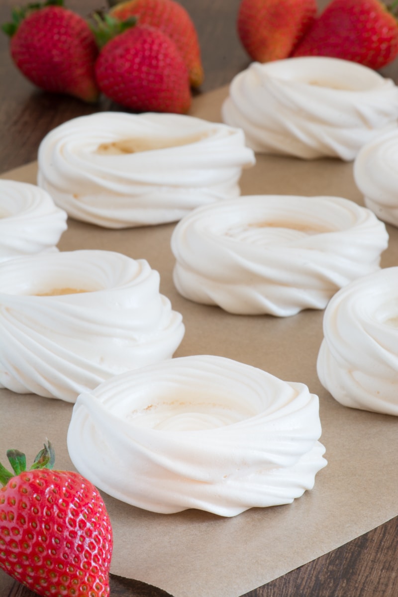 Meringue nests on a sheet of baking parchment with fresh strawberries in the background.