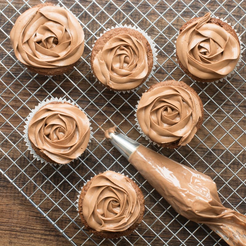 A piping bag full of Nutella buttercream surrounded by Nutella cupcakes topped with Nutella buttercream roses.