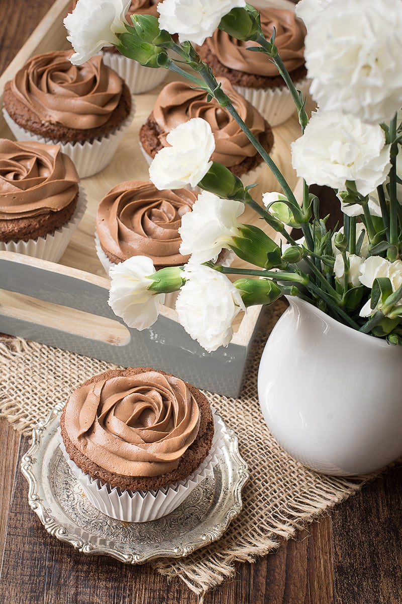 A Nutella cupcake topped with Nutella buttercream on a small silver dish next to a vase of white carnations. There's a tray of Nutella cupcakes in the background.