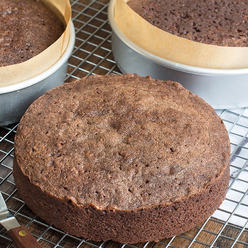 A layer of road chocolate cake on a cooling rack with two more chocolate cakes in tins in the background.