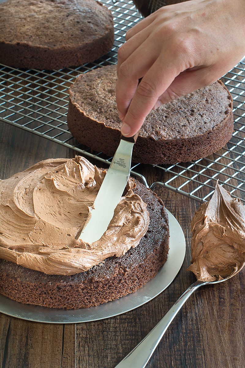 Spreading chocolate buttercream onto a round chocolate birthday cake using a palette knife. There are two more layers of chocolate cake on a cooling rack in the background.