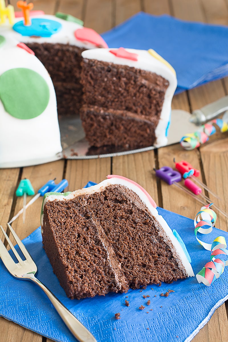 A slice of chocolate birthday cake filled with chocolate buttercream and covered in fondant icing on a blue napkin with a cake fork at the side. There are candles laying just behind it and behind that it the rest of the cake with another slice being removed on a cake slice.