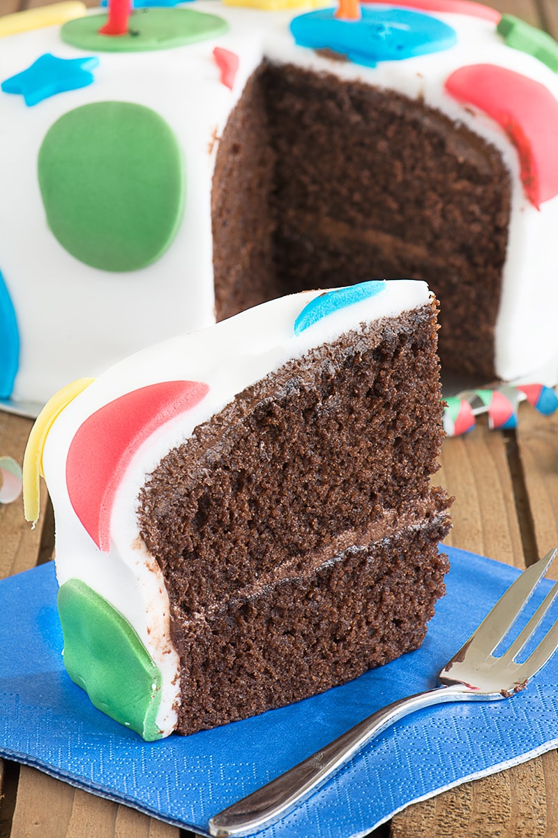 A slice of chocolate birthday cake filled with chocolate buttercream and covered with fondant icing. The rest of the cake is in the background with a gap showing where the slice had come from.