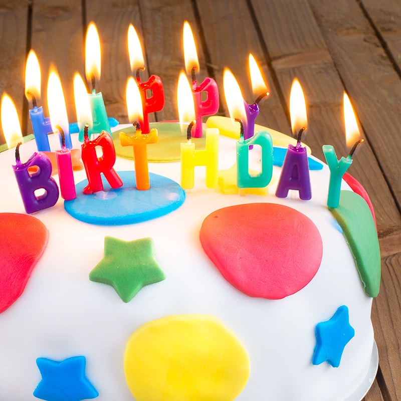 A round birthday cake covered in white fondant icing decorated with multi-coloured stars and circles. The cake is topped with burning candles spelling "HAPPY BIRTHDAY"