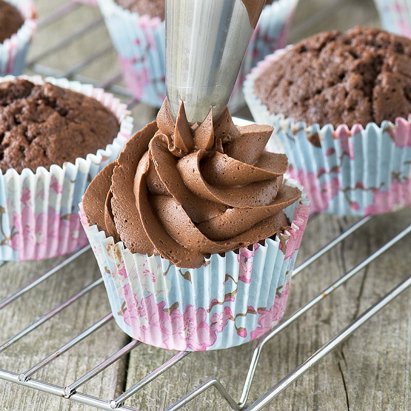 Piping chocolate buttercream onto a chocolate cupcake.