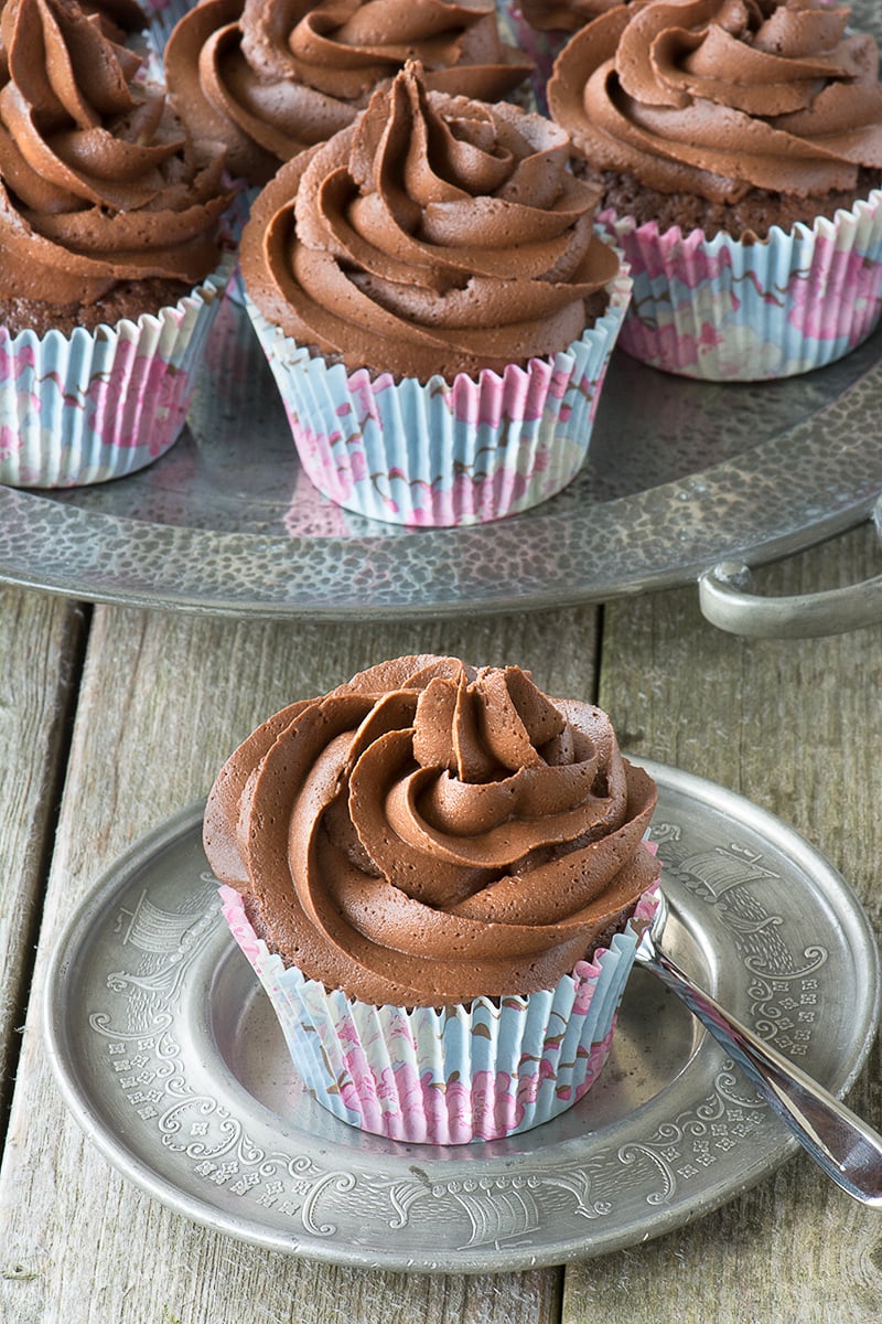 A chocolate cupcake on a round silver dish with a cake fork next to it. There are lots of chocolate cupcakes on a silver platter in the background.