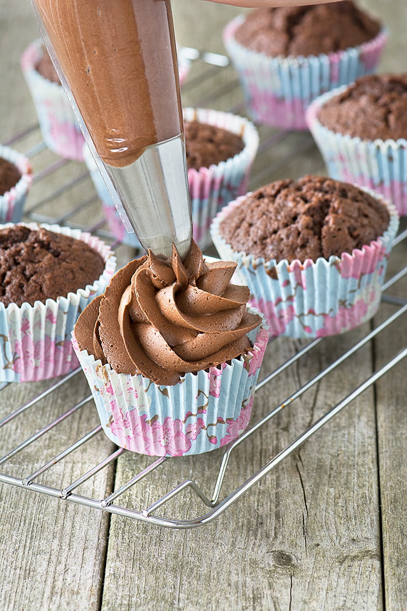 Piping chocolate buttercream onto a chocolate cupcake. There are several plain chocolate cupcakes in the background.