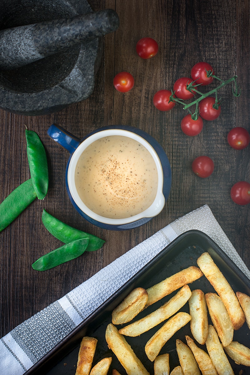 Looking down on a blue jug of peppercorn sauce with a baking tray of chunky chips, cherry tomatoes, mangetout and a dark grey pestle and mortar.
