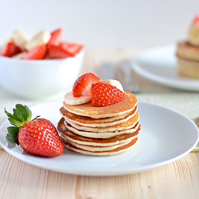 A stack of American pancakes topped with fresh fruit with a bowl of fruit and more pancakes in the background.
