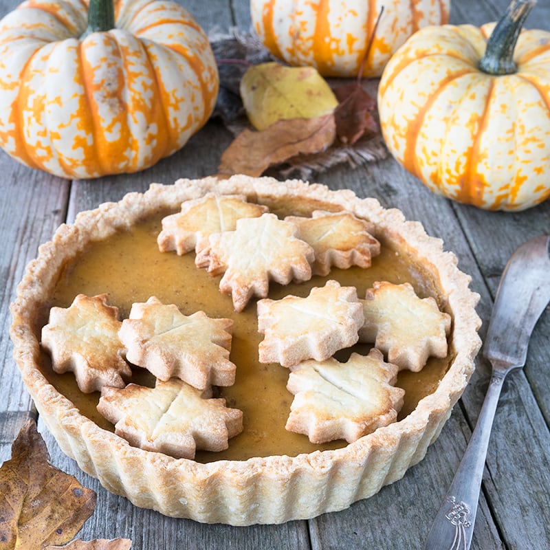 Angled view of a whole pumpkin pie decorated with pastry leaves on a wooden table with pumpkins in the background. 