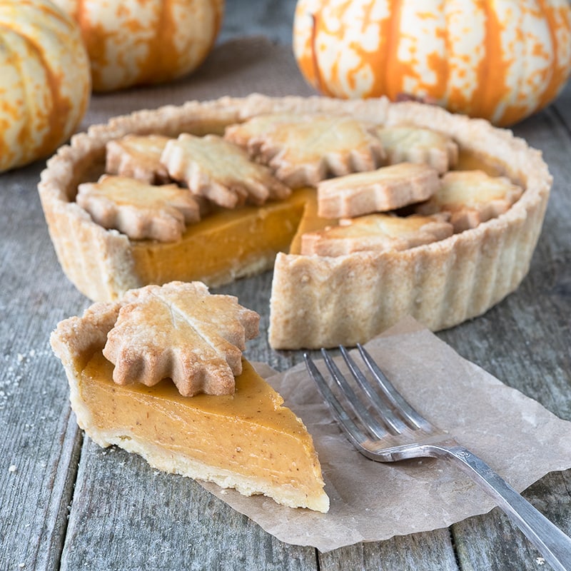 A slice of pumpkin pie with decoration shown in front of the rest of the pie and fresh/whole pumpkins. 