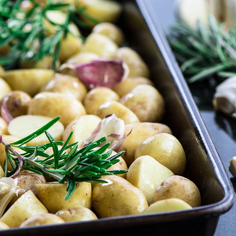 New potatoes in a baking tray ready to be roasted. They're tossed in olive oil and then topped with sprigs of rosemary and crushed cloves of garlic.