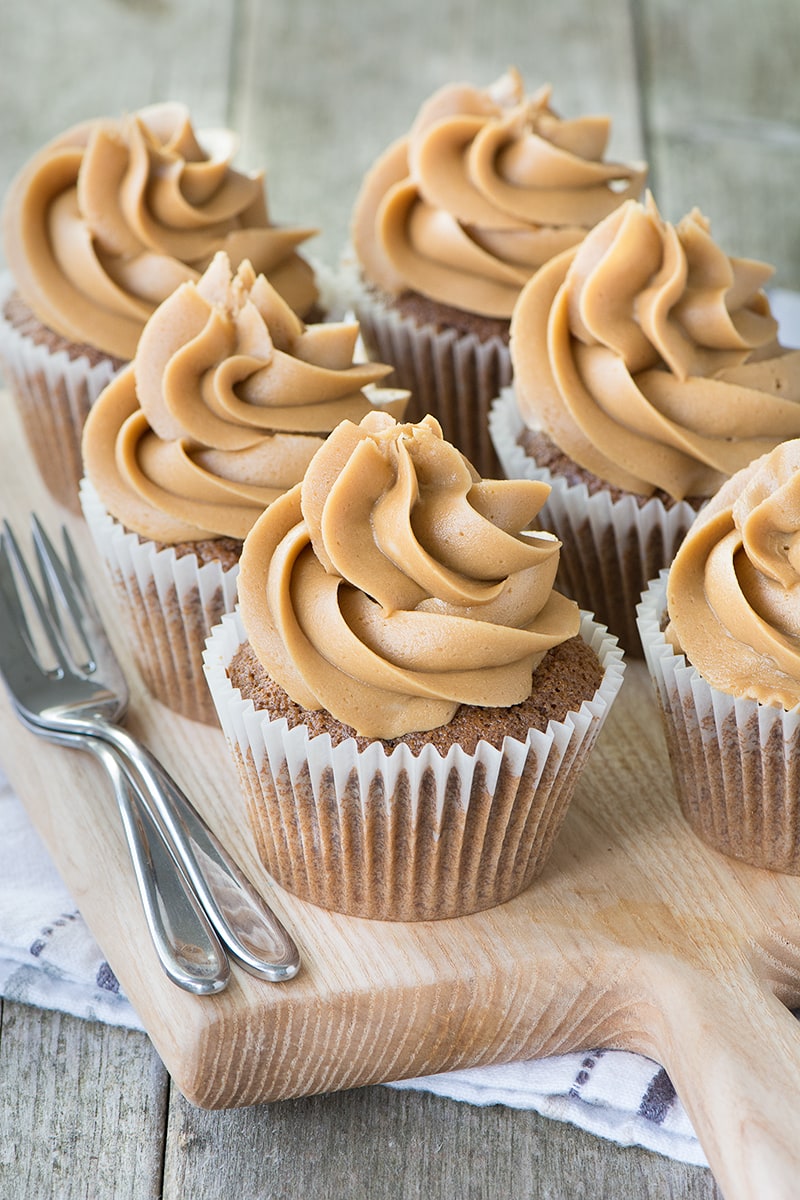 Six coffee cupcakes topped with coffee buttercream on a wooden board with cake forks at the side.