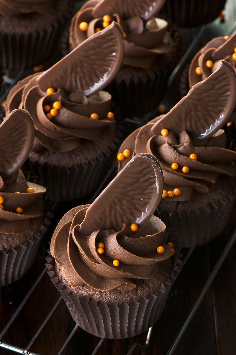 Chocolate orange cupcakes topped with chocolate orange buttercream, a slice of Chocolate Orange and orange sprinkles on a cooling rack.