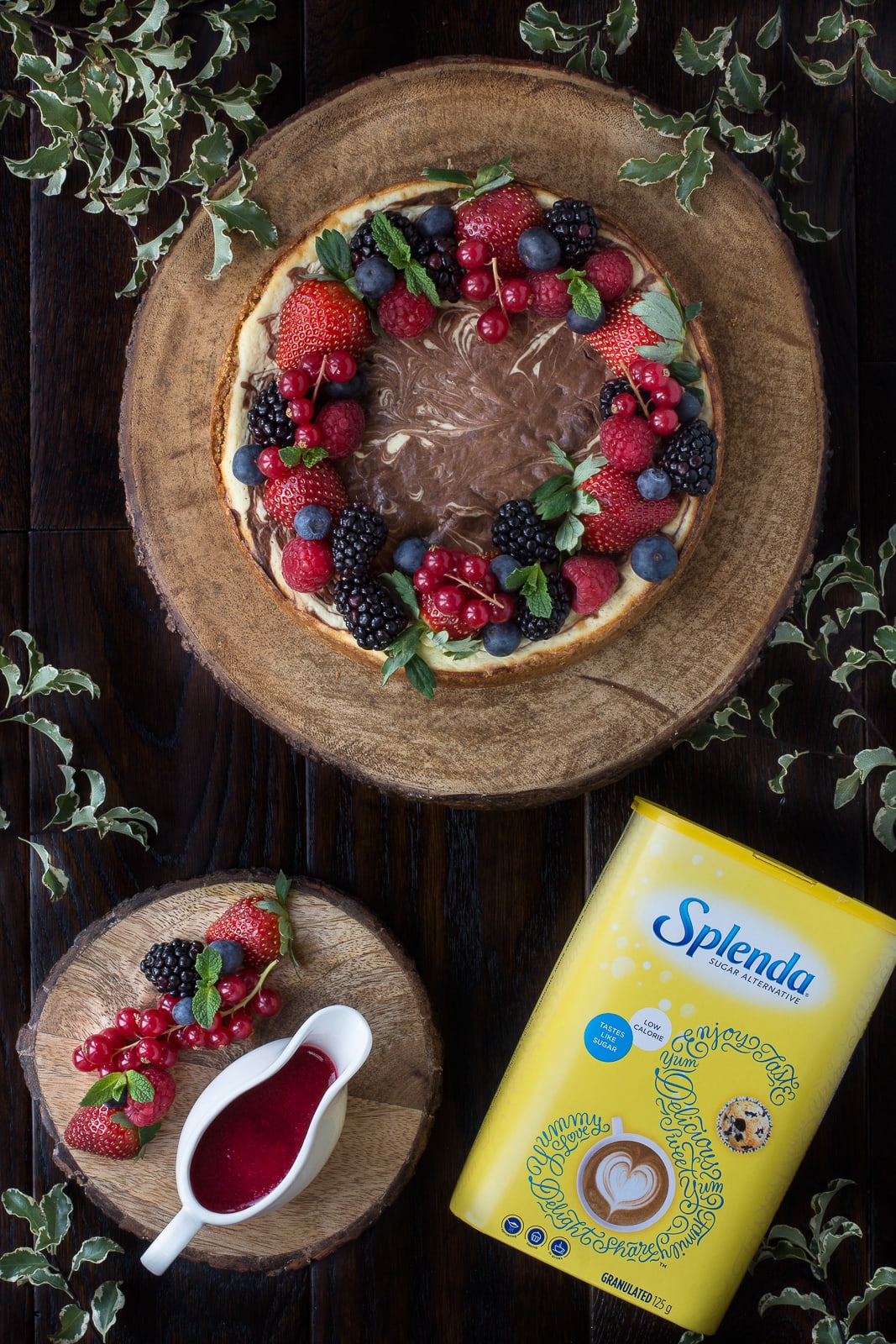 Looking down on a baked chocolate marbled cheesecake topped with fresh fruit on a wooden board. There's also a jug of recurrent coulis and a box of SPLENDA.