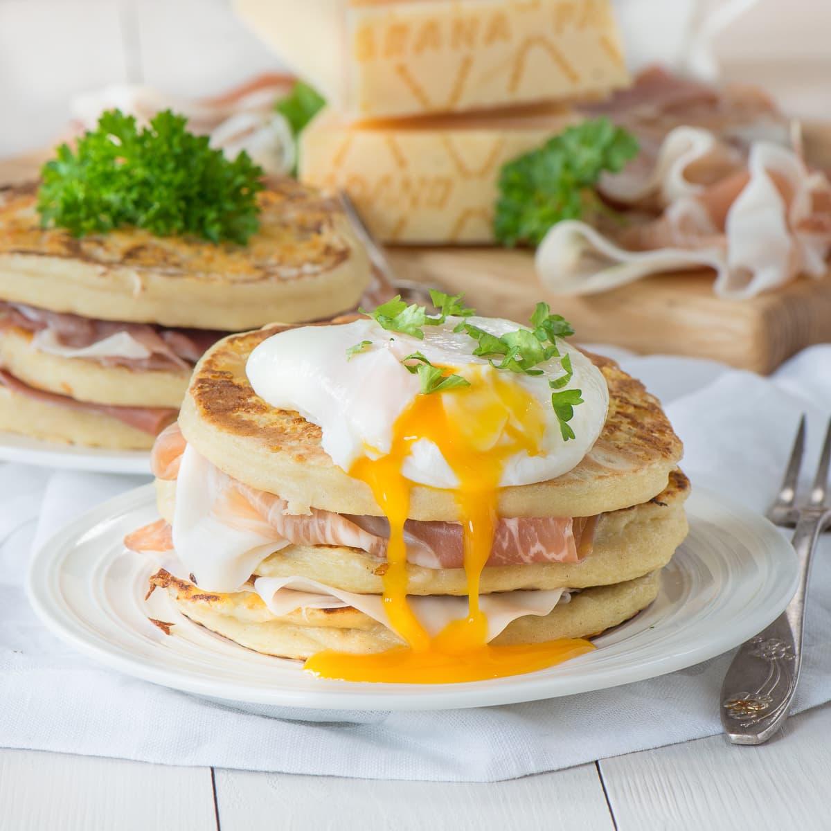 A stack of Grana Padano cheese pancakes, layered with Prosciutto di San Daniele and topped with a poached egg with the yolk running down the side.