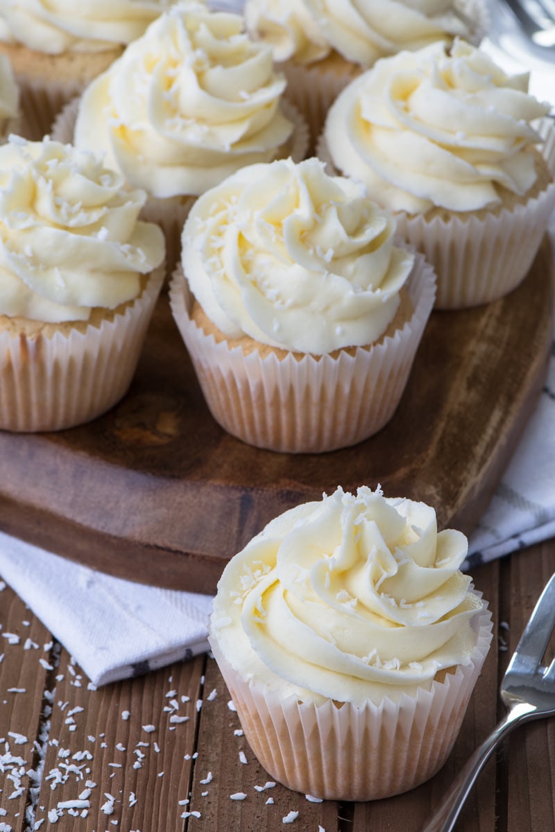 A coconut cupcake topped with coconut buttercream next to a cake fork with more cupcakes on a wooden board in the background.