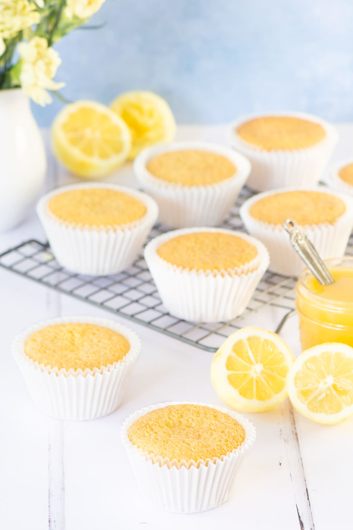 Lemon cupcakes without buttercream in white cupcake cases. Some on the table and some on a cooling rack. There are lemons, lemon curd and yellow flowers in the background.