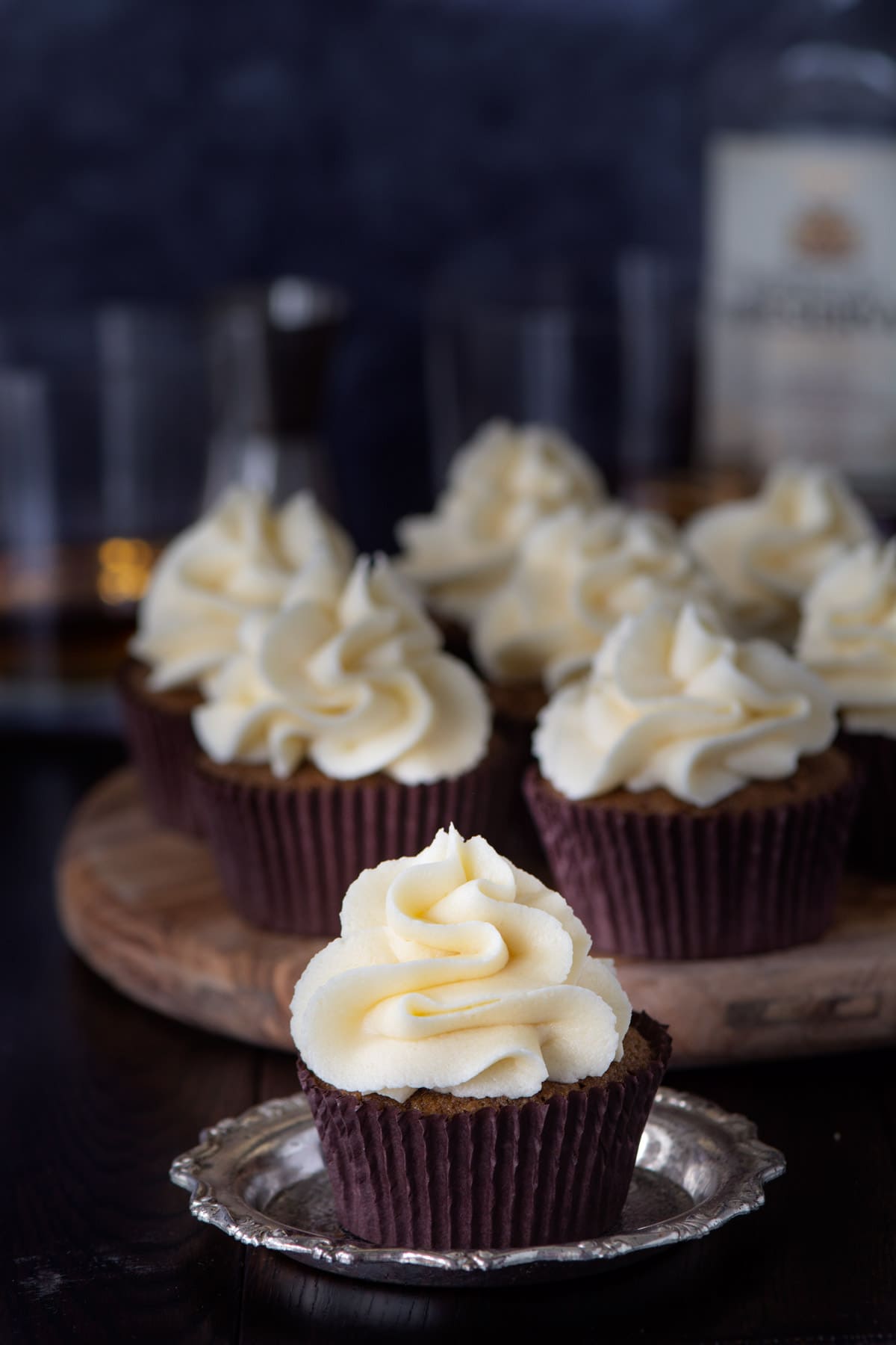 A coffee cupcake topped with whiskey buttercream. There are more cupcakes and a bottle and glasses of whiskey in the background.