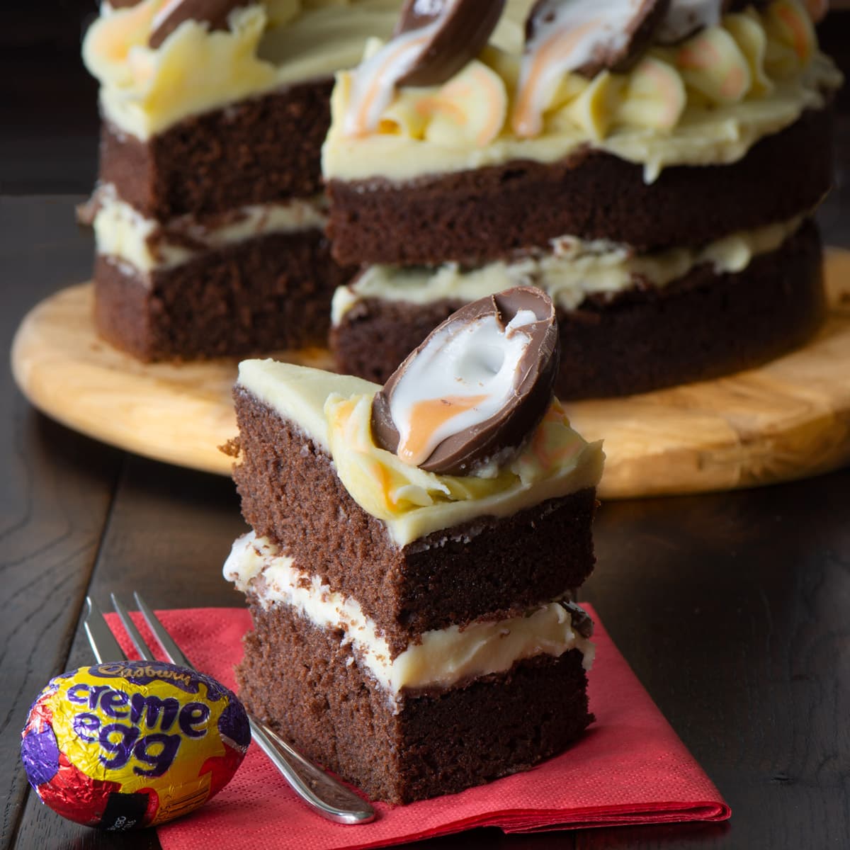 A slice of Creme Egg cake with the remaining cake in the background. A chocolate cake, with white chocolate buttercream, topped with Creme Egg halves.