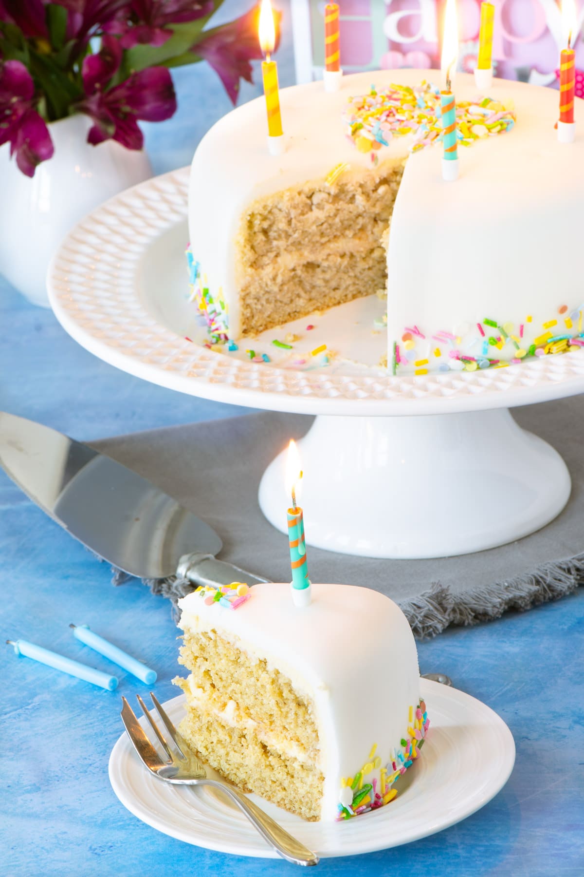 A slice of vegan birthday cake filled with vegan vanilla buttercream on a plate. The cake is decorated with vegan buttercream, white fondant icing and colourful sprinkles.