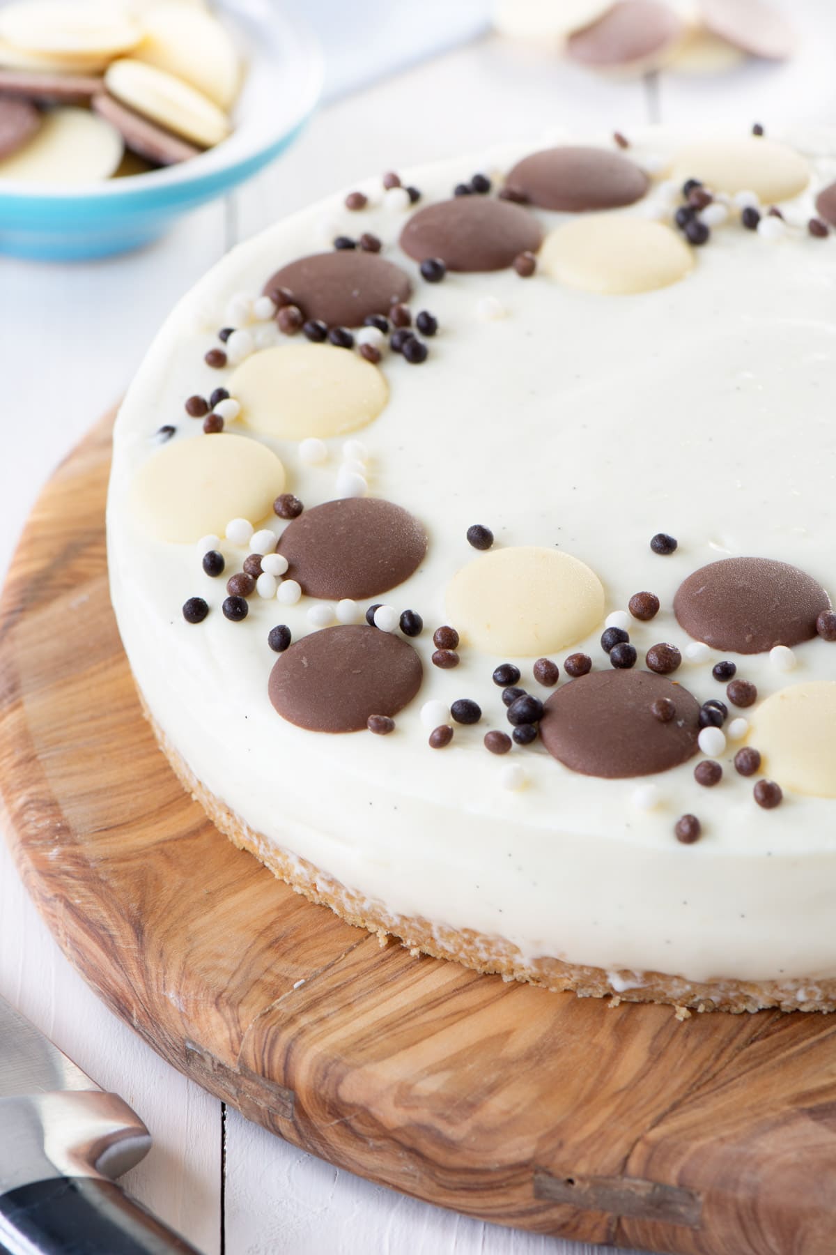 No-bake white chocolate cheesecake on a wooden board. The cheesecake is decorated with white and milk chocolate buttons and chocolate sprinkles.