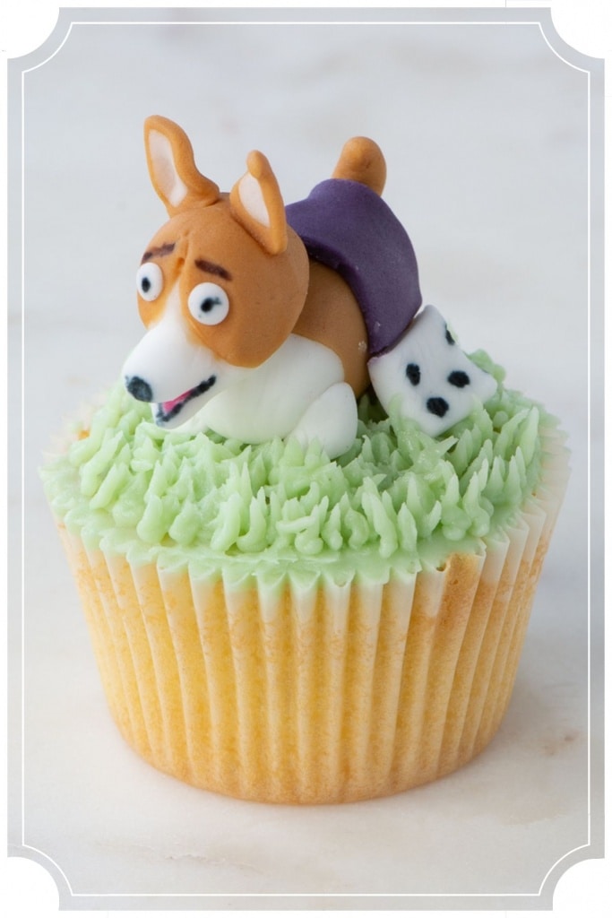 Cupcake topped with green buttercream grass and a fondant corgi with a purple regal robe.