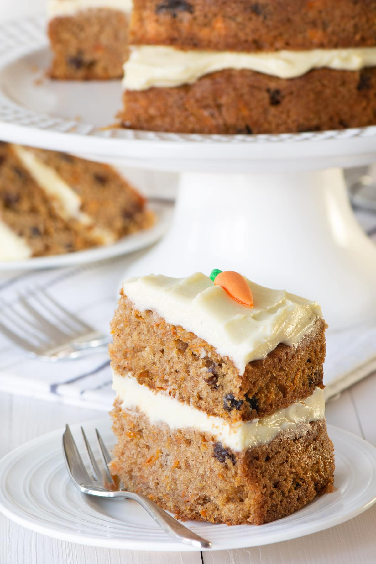 A slice of carrot cake on a white plate. The remaining cake is in the background. The slice shows the raisins running through the carrot cake sponge.
