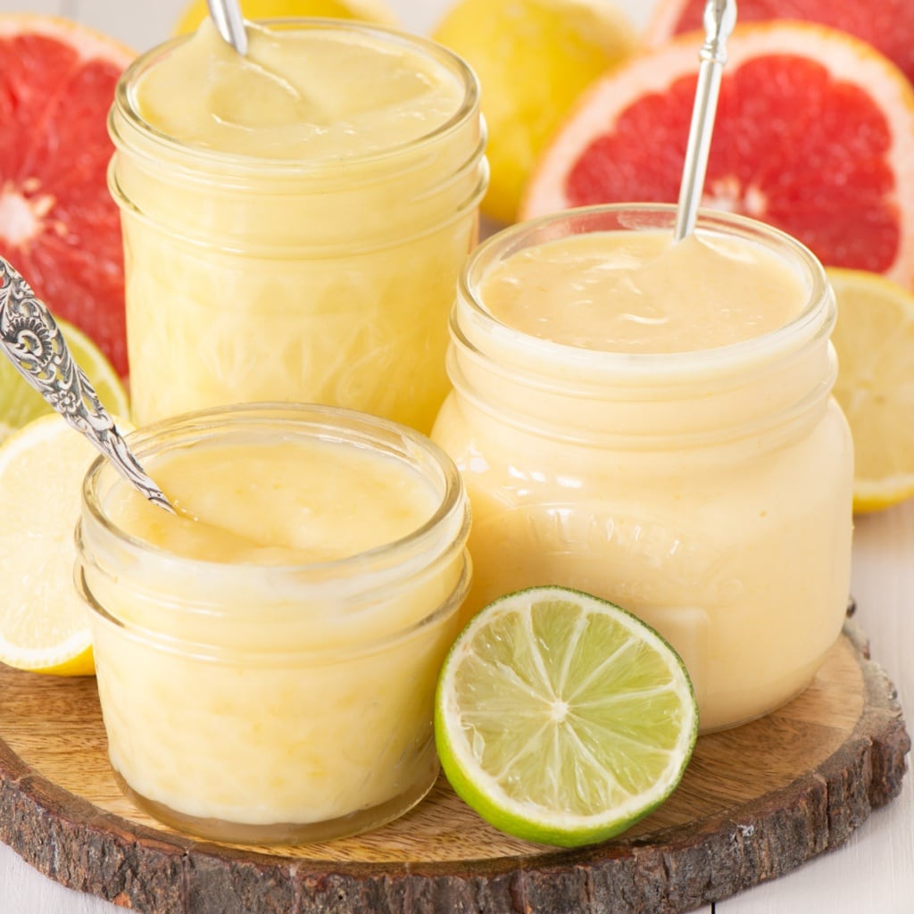 Three jars oh homemade curd - lemon, lime and grapefruit. The jars are on a wooden board and are surrounded by cut citrus fruit.