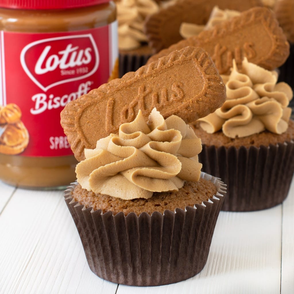 A Biscoff cupcake topped with Biscoff buttercream and a Biscoff biscuit. There is a jar of Biscoff spread and a packet of Biscoff biscuits in the background. Behind them are more decorated Biscoff cupcakes.