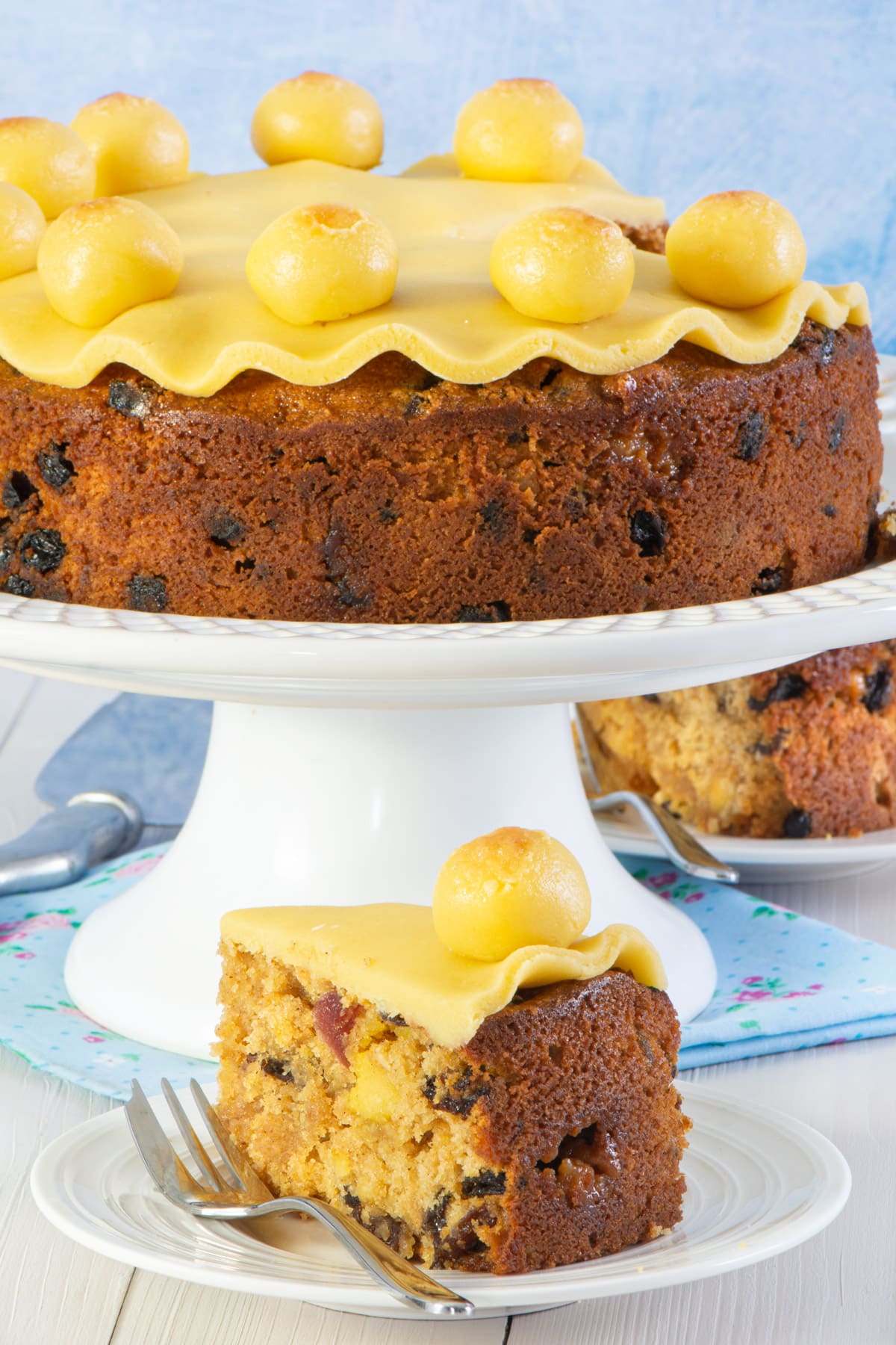 Slice of decorated simnel cake in front the the rest of the cake on a cake stand. 